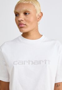 Fehér pamut póló, melynek mellkasán strasszos "Carhartt" logó található, kerek nyakkivágással és rövid ujjakkal.