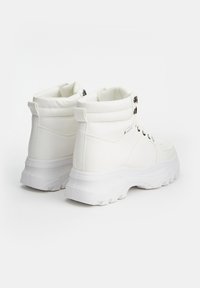 Botas blancas hasta el tobillo, hechas de material sintético, con una suela gruesa, un collar acolchado y ojales de metal para los cordones. Diseño de punta redonda.