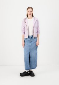 Giacca a zip lilla, t-shirt bianca e gonna maxi in denim blu con spacco frontale. Scarpe platform nere completano l'outfit.