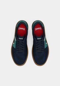 Pari tummansinisiä ja turkooseja nyörikenkiä, joissa on ruskeat pohjat ja punaiset pohjalliset, joissa lukee "Camper", kuvattu ylhäältä valkoisella taustalla.