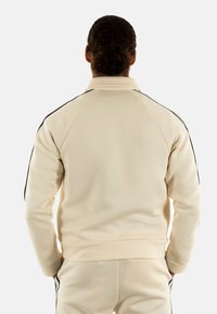 Sudadera beige con cuello alto, que presenta franjas laterales negras, una silueta ajustada y puños y bajo acanalados. Textura de tejido suave y liso.