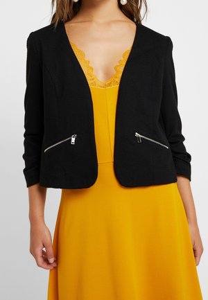 Frau trägt einen schwarzen strukturierten Blazer mit Reißverschlusstaschen über einem senfgelben Kleid mit Spitzenbesatz am Ausschnitt.