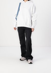 Sweat à capuche blanc avec une poche avant et un logo discret, associé à un jean noir à jambes larges et des baskets rose clair. Sangle d'épaule bleue en accent.