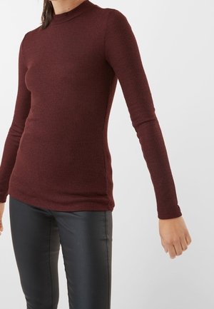 Femme portant un pull moulant côtelé bordeaux à manches longues et un pantalon noir, sur un fond clair uni.