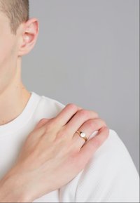 Zelta gredzens ar baltu akmeni, uz rokas, kas balstās uz baltas teksturētas auduma. Smalka spīdēšana un gluda apdare.