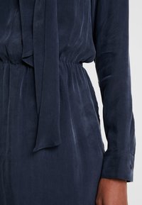 Abito blu scuro con colletto a cravatta, maniche lunghe e vita elastica. Tessuto liscio con una leggera lucentezza e dettaglio con bottoni sui polsini.