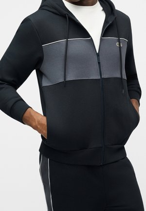 Mann trägt einen schwarzen Kapuzenpullover mit Reißverschluss und grauer Brustpartie, die Hände in den Taschen, kombiniert mit schwarzen Hosen mit weißen Seitenstreifen.