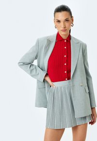 Grijze blazer met twee knopen, rode kabelgebreide trui en grijze geplooide minirok. Gladde stof, taillering en gestructureerd ontwerp.