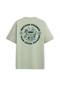 Ανοιχτό πράσινο T-shirt με σκούρο πράσινο καρτούν φλιτζάνι τσαγιού matcha που κρατά ένα κουτάλι, περιτριγυρισμένο από κείμενο και εικονίδια σχετικά με το τσάι στην πλάτη.