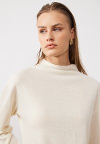 Tissu léger de couleur crème avec un design à col haut. Texture lisse avec une coupe décontractée. Boucles d'oreilles florales dorées ajoutent du contraste.