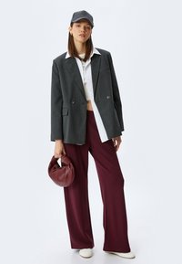 Blazer gris, camisa blanca, pantalones de pierna ancha burdeos y un bolso burdeos. La modelo lleva una gorra gris y zapatos blancos. Textura suave.