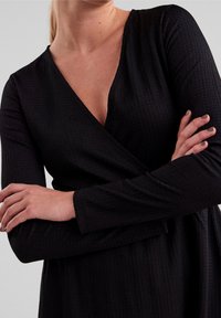 Robe noire à manches longues, en tissu texturé, avec un décolleté en V profond et des détails froncés au niveau de la taille. Matière douce et fluide.