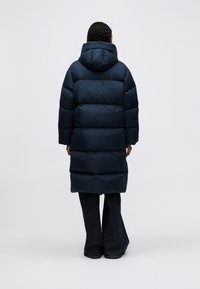 Osoba v dlouhém, tmavě modrém péřovém kabátu s kapucí, stojící zády k nám na jednoduchém světle šedém pozadí.