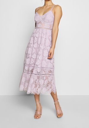 Femme portant une robe midi en dentelle lavande sans manches avec fines bretelles et sandales à talons nus, debout devant un fond uni.