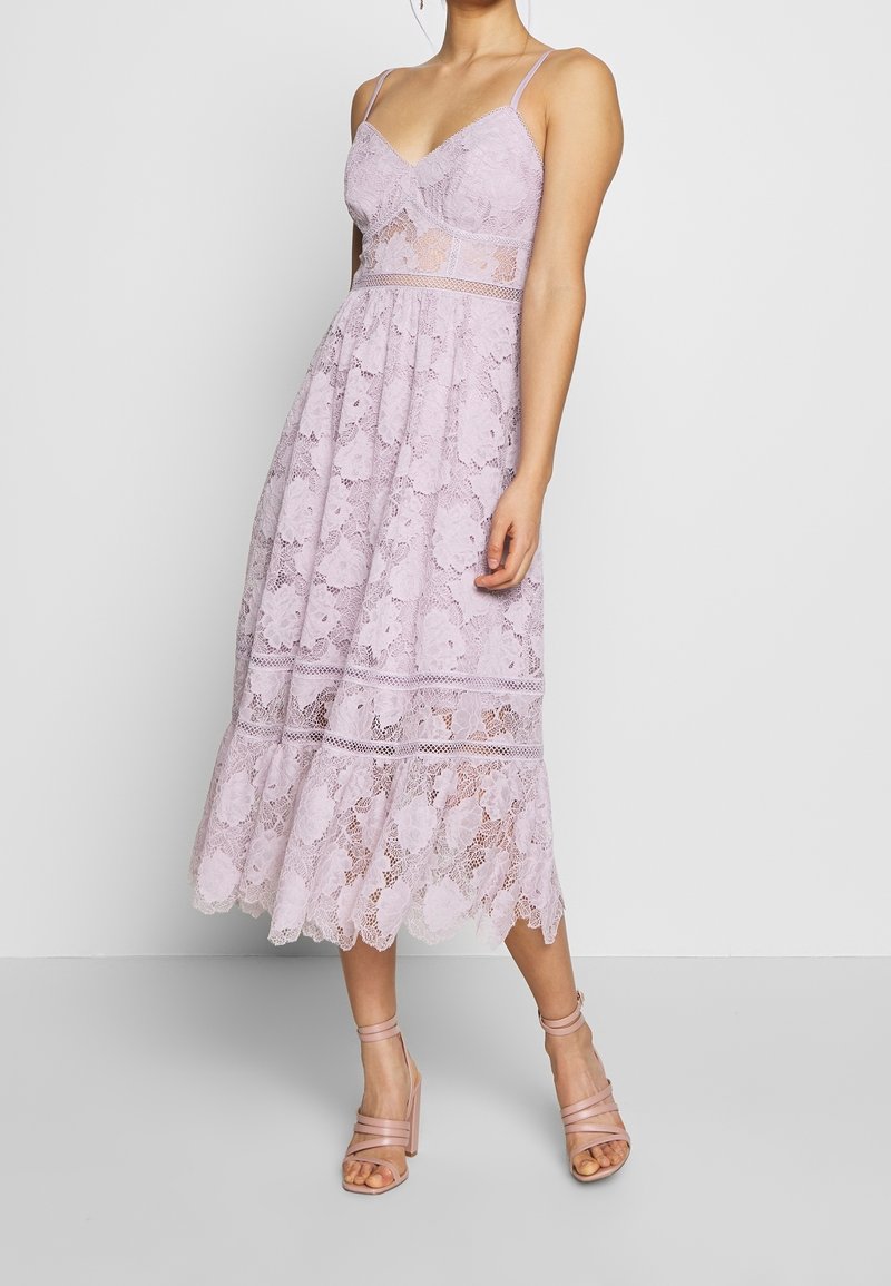 Femme portant une robe midi en dentelle lavande sans manches avec fines bretelles et sandales à talons nus, debout devant un fond uni.