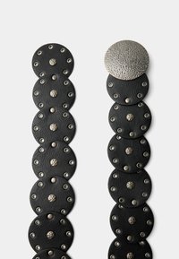 Svart läderbälte med cirkulära länkar och silverfärgade metalldekorationer samt en texturerad cirkulär spänne. Platt design med släta och präglade texturer.