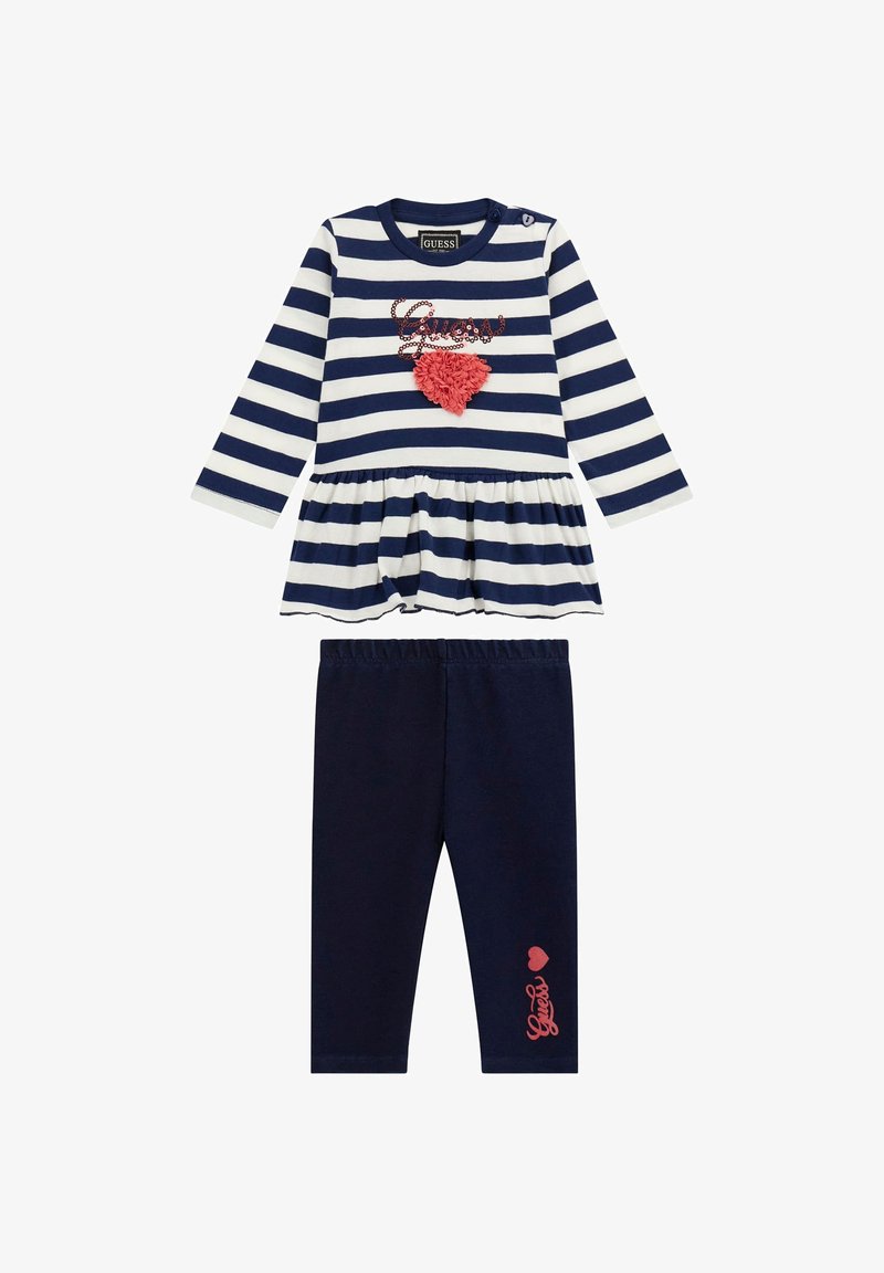 Csíkozott sötétkék és fehér peplum felső korall szív részlettel, sötétkék leggingsszel párosítva, amelyen korall színű "Guess" felirat található a lábon.