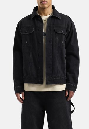 Zwarte denimjack met een klassieke kraag, voorzakken en knoopsluiting. Draagt over een lichtbeige t-shirt met lange mouwen en een ontspannen pasvorm.