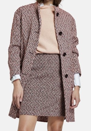 Femme portant une veste et une jupe en tweed coordonnées dans des tons marron et blanc avec un pull en tricot beige et des poignets de chemise blancs visibles.