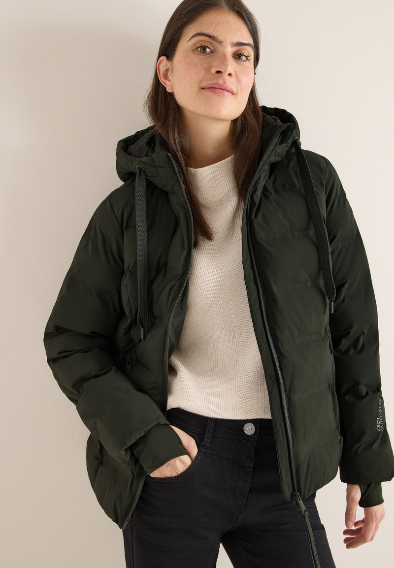 Giacca a palloncino verde scuro con cappuccio e cordini regolabili, dotata di chiusura con zip e un maglione beige chiaro sottostante.