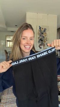 Lächelnde Frau hält eine schwarze Karl Lagerfeld Jeans am Bund in einem modernen, gut beleuchteten Wohnzimmer hoch.