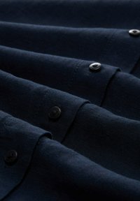 Tessuto blu navy con una superficie liscia, caratterizzato da diversi bottoni neri lungo i bordi e le cuciture, disposti in pieghe sovrapposte.