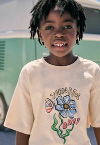 Beige t-shirt med korte ærmer med et trykt blåt blomsterdesign, en sommerfugl og teksten "VIDUNDERLIGE DAGE" i sort skrift.