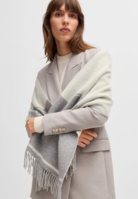 Châle gris et crème avec des bords frangés, drapé sur un blazer gris clair avec trois accents de boutons. Texture douce, design superposé.