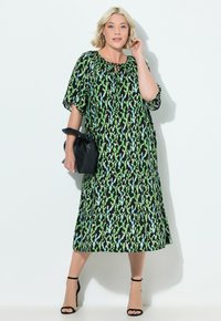 Schwarzes Kleid mit grünem und blauem Blumenmuster, kurzen schwingenden Ärmeln, rundem Ausschnitt, gestuften Saum, kombiniert mit einer schwarzen Handtasche und Riemchensandalen.