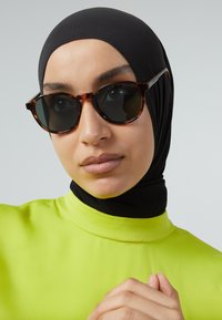Occhiali da sole tartarugati con lenti scure, forma rotonda e accessori neri eleganti. Il modello indossa un top verde brillante e un hijab nero.