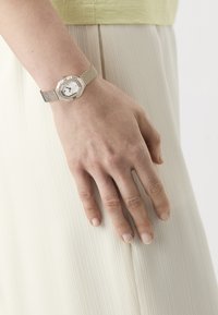 Octagonal silver-tone watch with a mesh band, white face, small black hands, and clear rhinestone accents, worn on a hand resting on a pleated skirt.