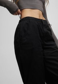 Pantaloni neri con vita elastica e tasche laterali, realizzati in tessuto liscio. Presentano un sistema di regolazione con lacci e un design dal taglio comodo.