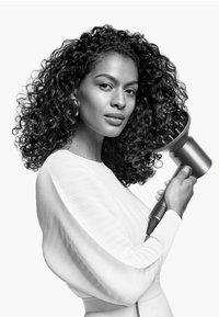 Haardroger met een metallic afwerking en een diffuseropzetstuk, vastgehouden door een persoon in een textuurrijke witte top, met zachte krullen en een natuurlijke huidtoon.