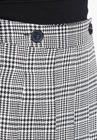 Sukně v černobílém houndstooth vzoru, která má knoflíkový detail v pase a texturovanou látku. Výrazný vzor s propracovaným střihem.