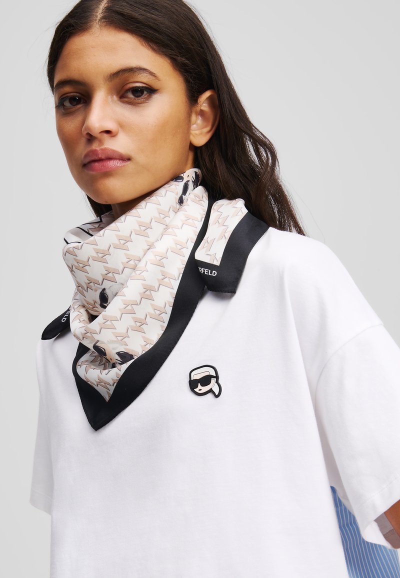 Weißes Baumwoll-T-Shirt mit rundem Halsausschnitt, ausgestattet mit einem schwarzen und beigen gemusterten Seidenschal und einem kleinen schwarzen Pin-Akzent auf der Brust.