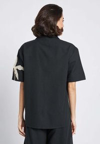 Maglietta nera a maniche corte con una vista posteriore che mostra un design floreale in pizzo beige testurizzato sulla manica sinistra e una vestibilità comoda.