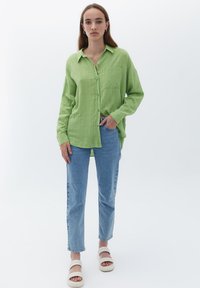 Jasnozielona koszula na guziki z teksturowanej tkaniny, z jedną kieszenią na piersi, podwiniętymi rękawami, w zestawieniu z jasnoniebieskimi prostymi dżinsami i beżowymi sandałami.
