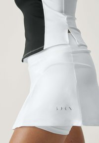 Falda deportiva blanca con un diseño de abertura, que cuenta con una parte superior negra. Material suave y elástico con un sutil detalle del logo en el dobladillo inferior.