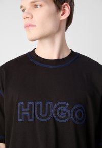 Melna kokvilnas T-krekls ar zilu izšūtu "HUGO" logo, noapaļota kakla izgriezums ar zilām akcentu stitched, īsām piedurknēm un brīvu piegriezumu.