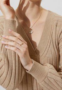 Pull en tricot côtelé beige avec un col en V, mettant en valeur un tissu texturé et des manches longues. Un collier chaîne doré avec un pendentif rond et une bague dorée à la main.