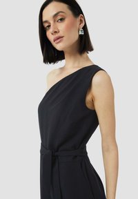 Zwarte one-shoulder jumpsuit met een aansluitende top, zijzakken en een knoopdetail in de taille, die de gladde stof en de gestructureerde silhouette toont.