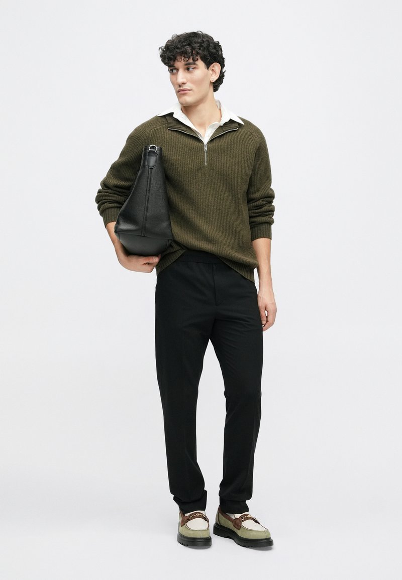 Groene geribbelde trui met daaronder een witte blouse, zwarte broek en een zwarte tas. De schoenen zijn multifunctioneel met een dikke zool.