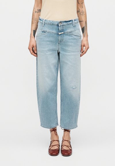 Jeans en denim bleu clair à coupe décontractée, avec une pièce usée au niveau du genou et une taille haute. L'ourlet est raccourci, assorti à des chaussures rouges à lacets.
