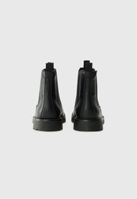 Botas de tobillo de cuero negro con una textura suave, lengüetas en la parte posterior y una suela de goma gruesa. La vista trasera muestra detalles de costura.
