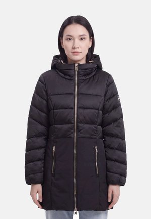 Donna che indossa un lungo giubbotto nero imbottito con cappuccio, cerniera frontale e tasche con cerniera, in piedi davanti a uno sfondo chiaro uniforme.