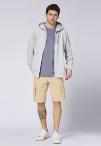 Gray zip-up hoodie, light blue t-shirt, tan cargo shorts, and white sneakers. Notable tattoo on leg and front-facing pose.