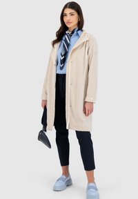 Cappotto impermeabile beige con zip, cappuccio con cordoncino, indossato sopra una camicia azzurro chiaro e pantaloni neri. Mocassini azzurro chiaro e una borsa completano il look.