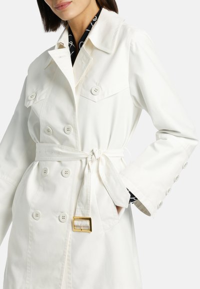 Manteau trench blanc à double boutonnage avec ceinture à nouer, grands boutons et poches à rabat. Présente des manches larges et un tissu lisse et léger.