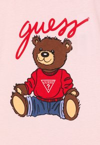 T-shirt rose clair avec un ours en cartoon au pelage marron, portant un pull rouge avec un logo blanc et un jean bleu. L'ours est assis.