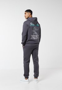 Uomo di spalle che indossa una felpa con cappuccio e pantaloni della tuta grigio carbone con testo grafico sulla schiena, in piedi su un pavimento bianco contro una parete bianca.